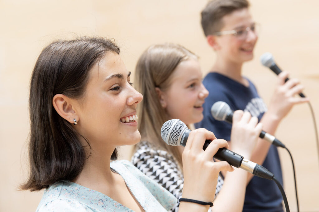 Zwei Mädchen und ein Junge singen mit Mikrophonen in der Hand.
